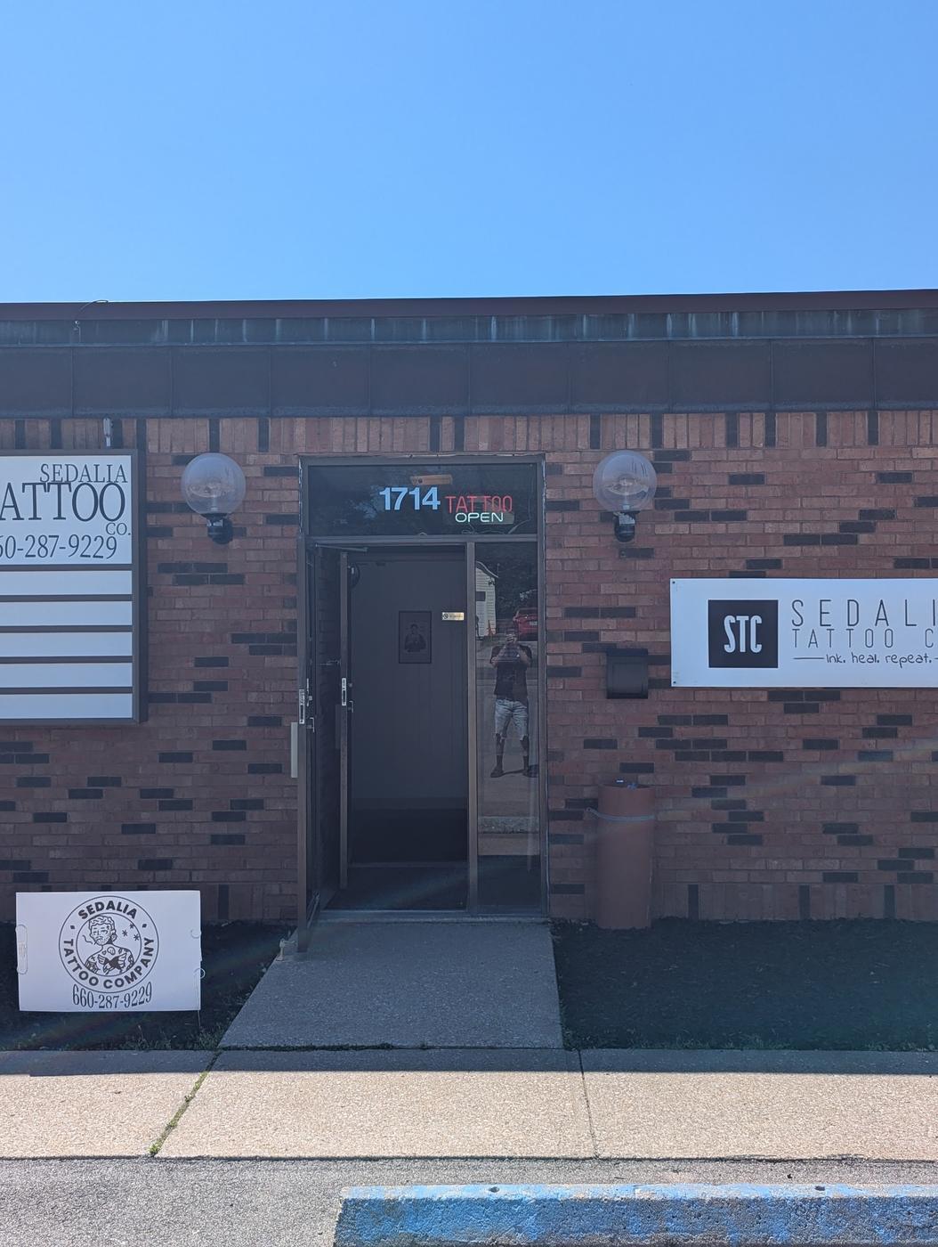 The Sedalia Tattoo Company shopfront: a brick unit with a dark door numbered 1714, exterior signage reading Sedalia Tattoo with the phone number, and a window decal.