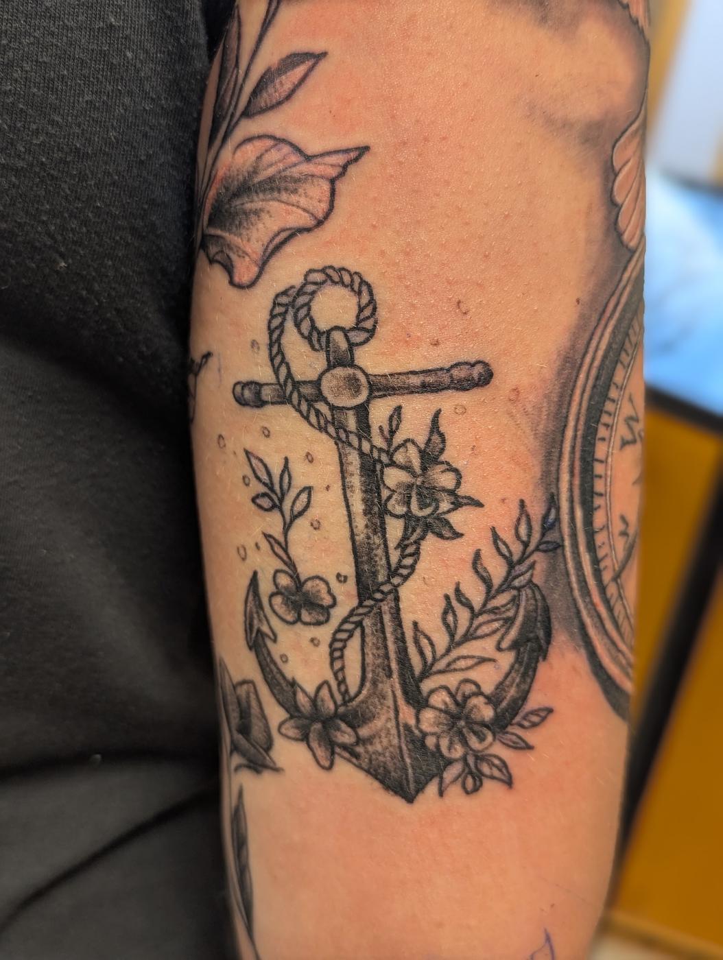 An anchor wrapped in flowers, tattooed in black and gray ink on an upper arm.