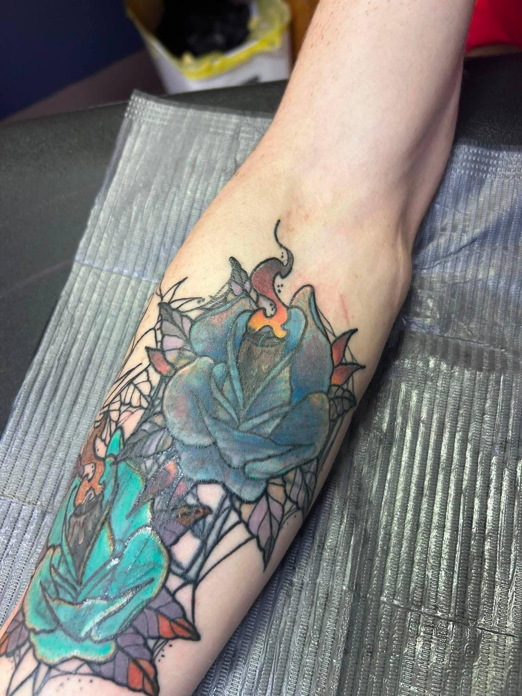Turquoise roses and a lit candle tattooed in color on a forearm, over fine web line work.