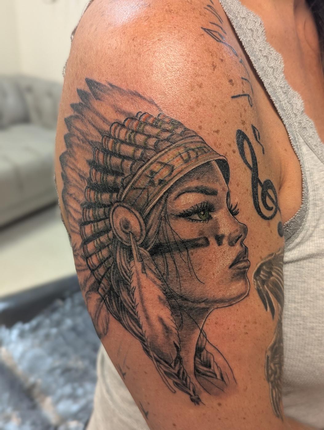 A portrait in a feathered headdress tattooed in black and gray ink on a shoulder.