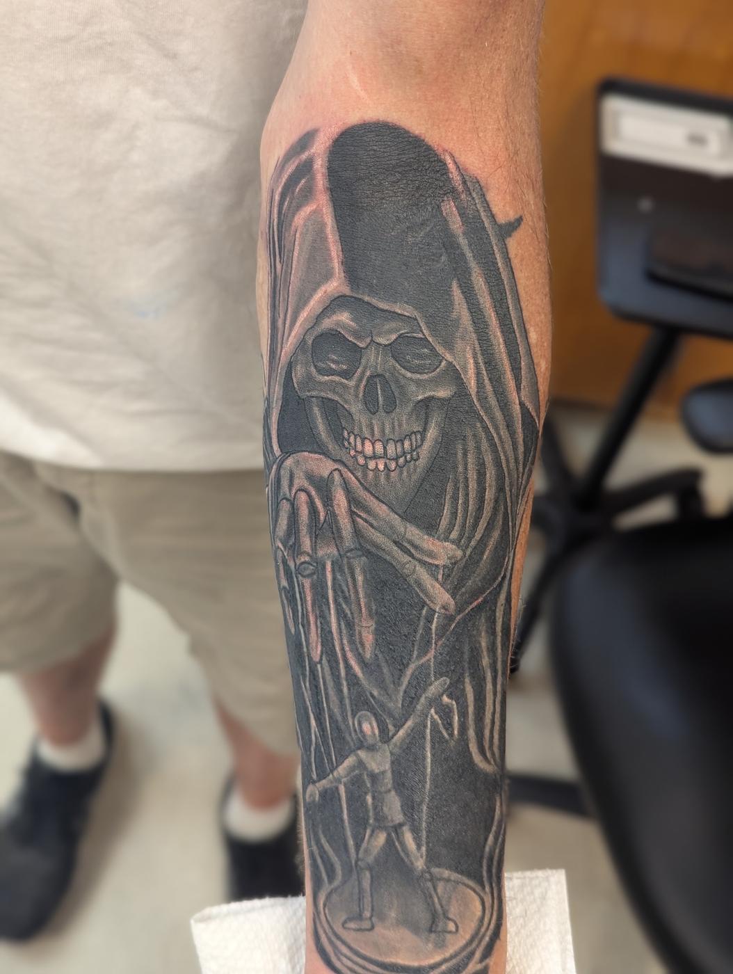 A hooded reaper with a skull face and skeletal hands, tattooed in black and gray ink on a forearm.